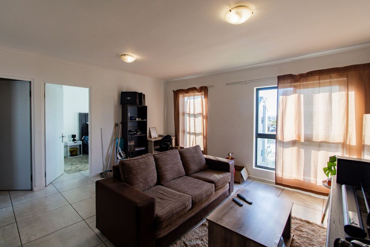 A bright living room featuring tiled flooring, large windows with natural light, and direct access to an adjacent bedroom.