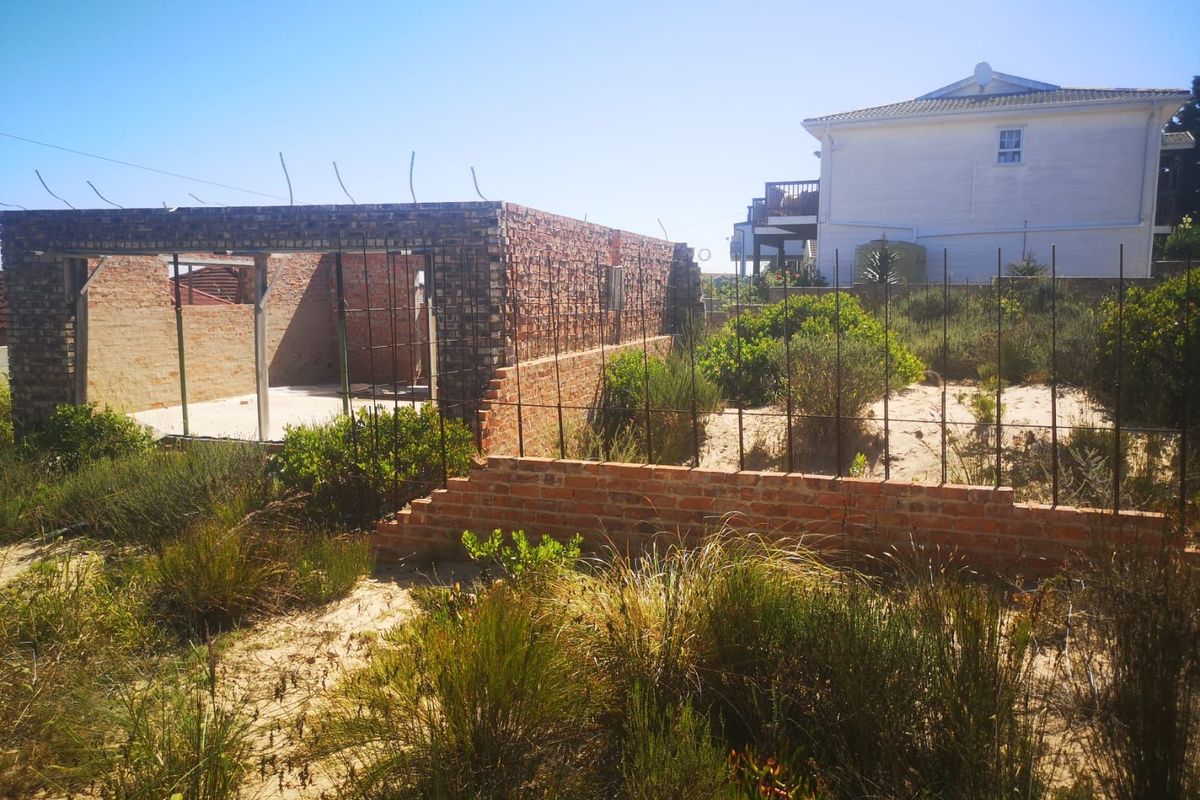 A vacant residential plot featuring an incomplete brick structure with exposed rebar, situated in a coastal or sandy environment.