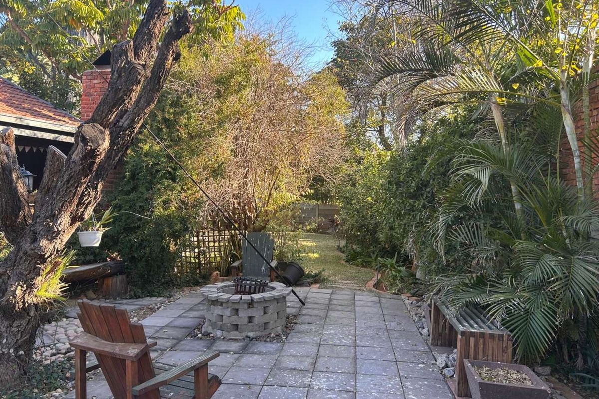 A private rear garden featuring a paved patio area with a built-in brick fire pit, surrounded by mature trees and established landscaping.