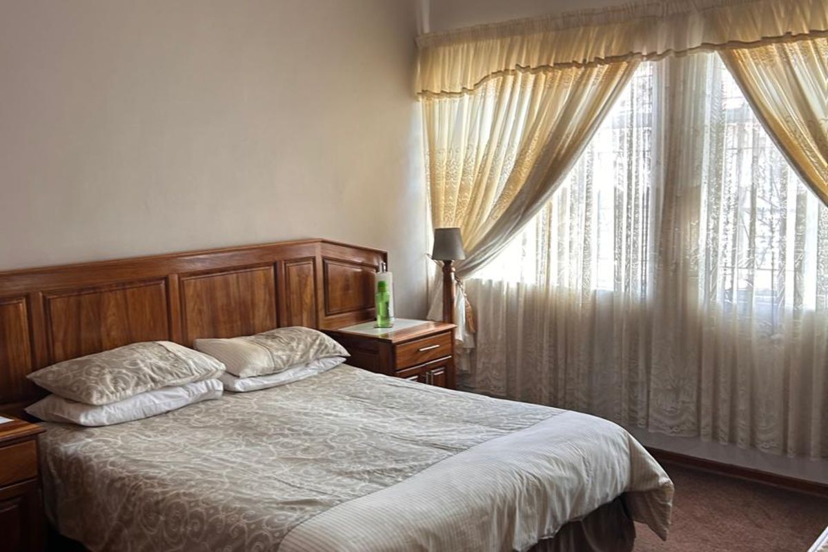 A comfortable bedroom featuring a wall-mounted air conditioning unit, carpeted flooring, and large windows with fitted curtains.