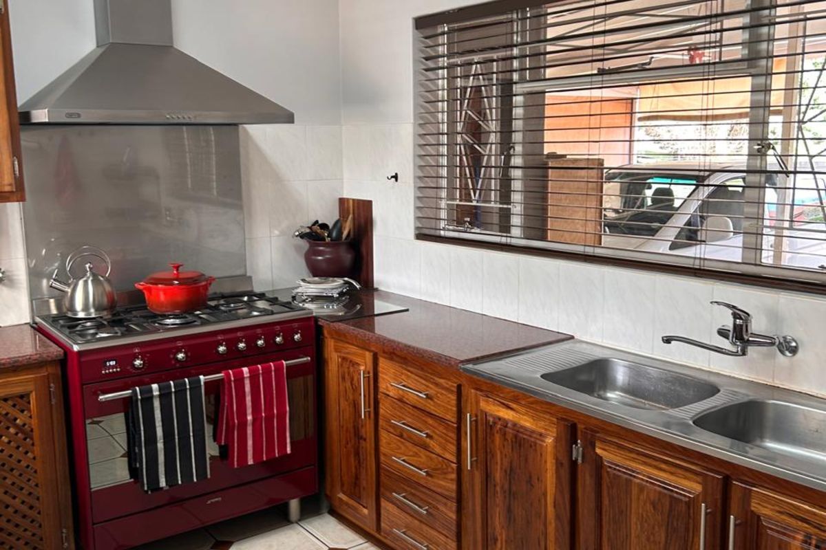 A functional kitchen featuring wooden cabinetry, a stainless steel double sink, a red freestanding gas stove with extractor fan, and tiled flooring.