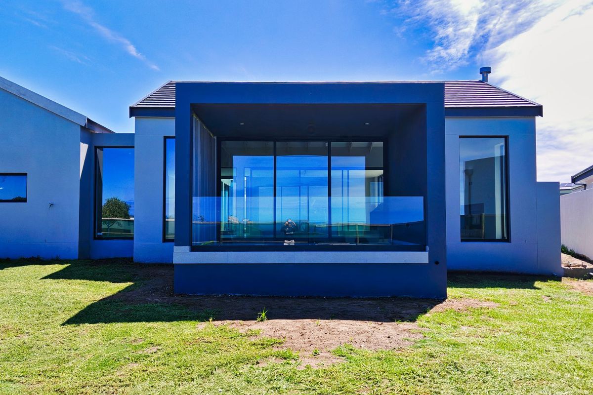 Modern rear exterior view of the house featuring a covered patio with glass balustrades, large floor-to-ceiling windows, and a well-maintained lawn.