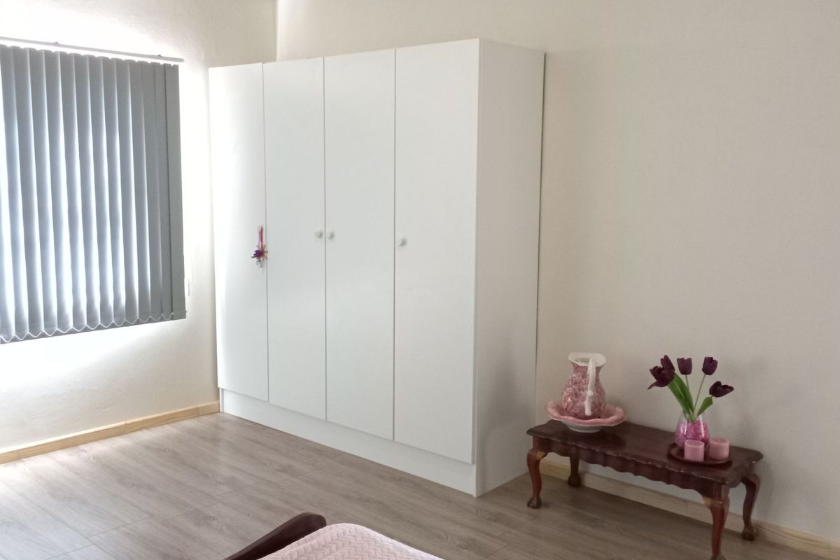 A bright bedroom featuring light wood-look laminate flooring, white built-in cupboards, and vertical window blinds.