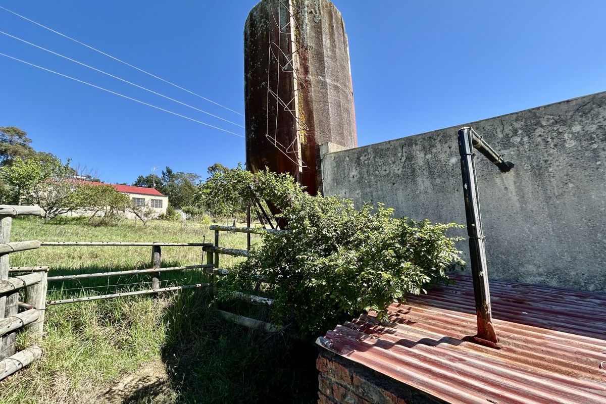 Exterior view of a farm property featuring a large metal silo, a weathered concrete boundary wall, and a rustic corrugated metal structure.