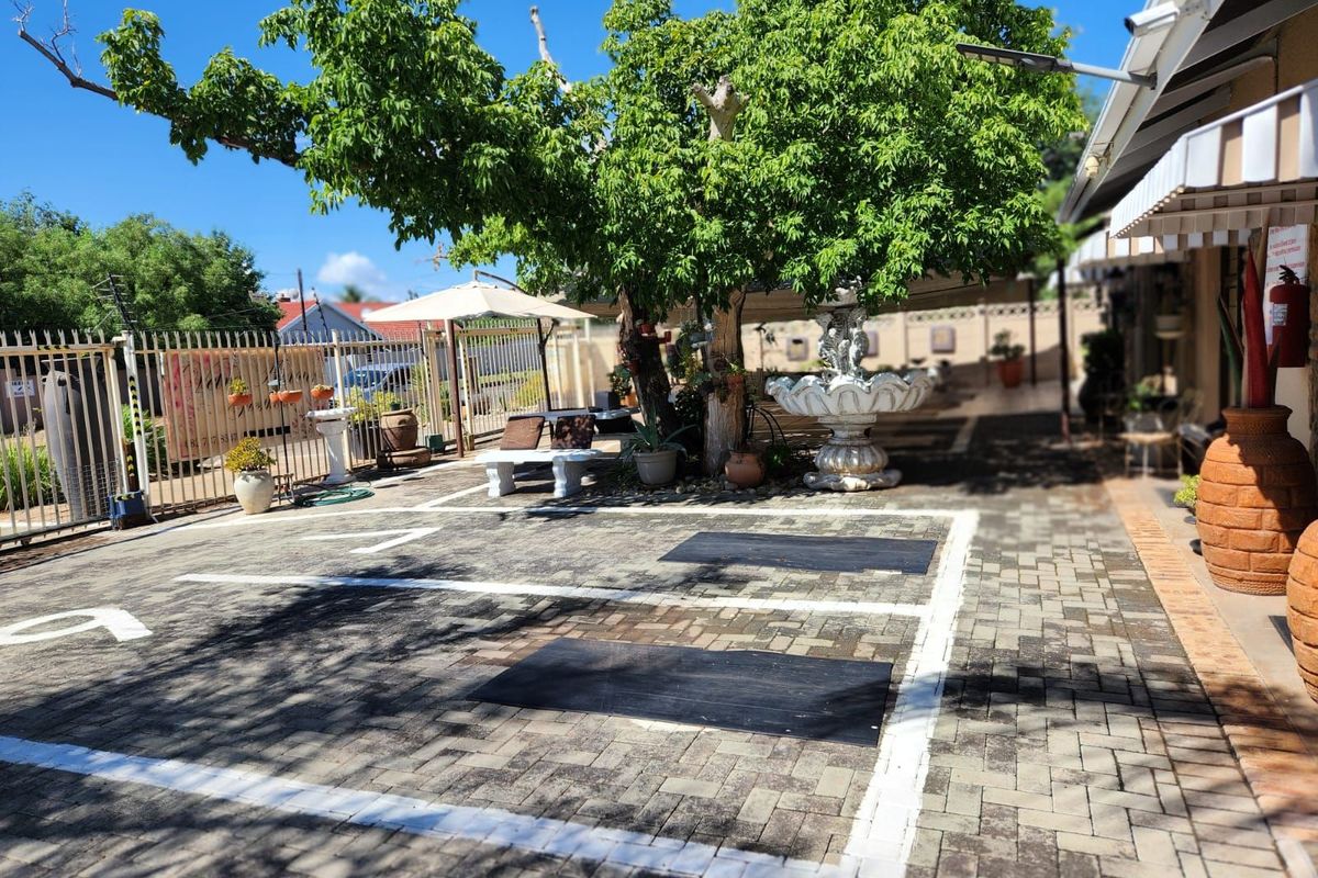 A paved outdoor courtyard area featuring designated parking bays, a large mature tree, and a decorative fountain, adjacent to the guest house building.