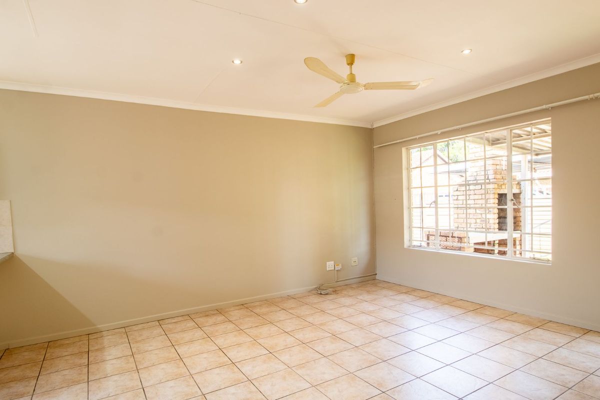 A bright, empty living area featuring neutral wall tones, tiled flooring, a ceiling fan, and large windows overlooking an outdoor brick braai area.