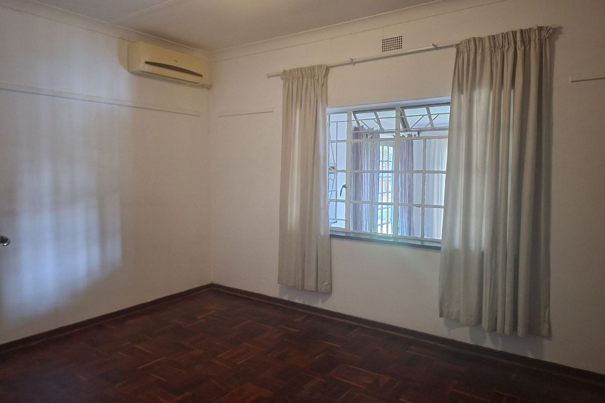 A bright, empty bedroom featuring polished parquet flooring, a wall-mounted air conditioning unit, and a window with security bars.