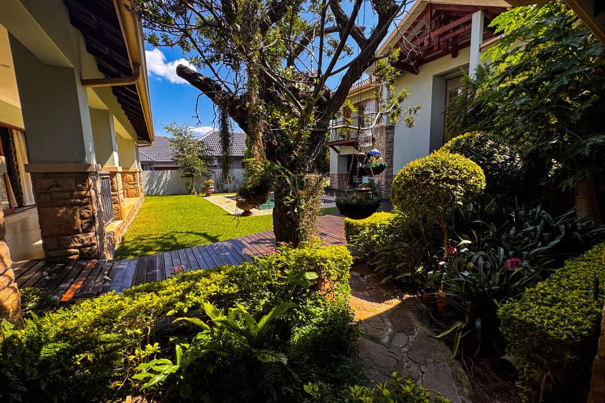 A lush, landscaped rear garden featuring a wooden deck, stone pathway, mature trees, and well-maintained shrubbery between two residential building structures.