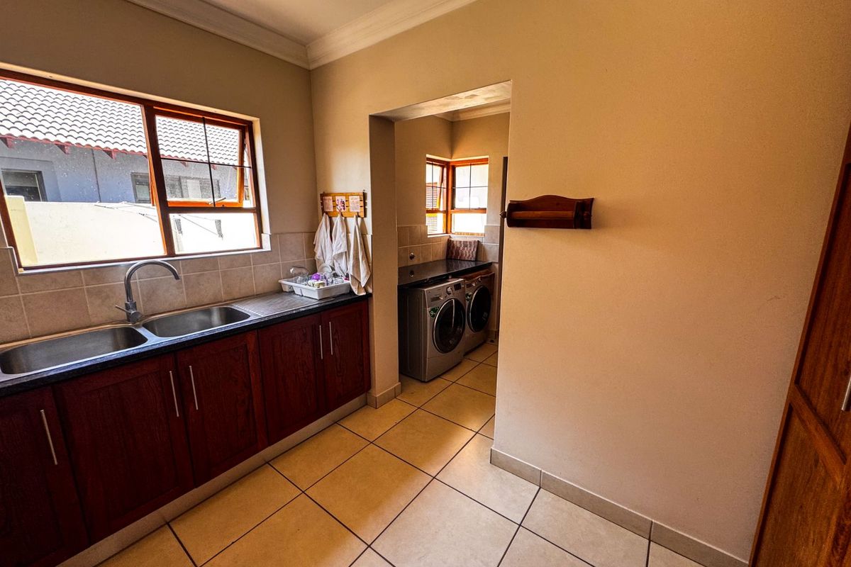 A functional kitchen featuring dark wood cabinetry, granite countertops, a double stainless steel sink, and an adjoining scullery area with tiled flooring.