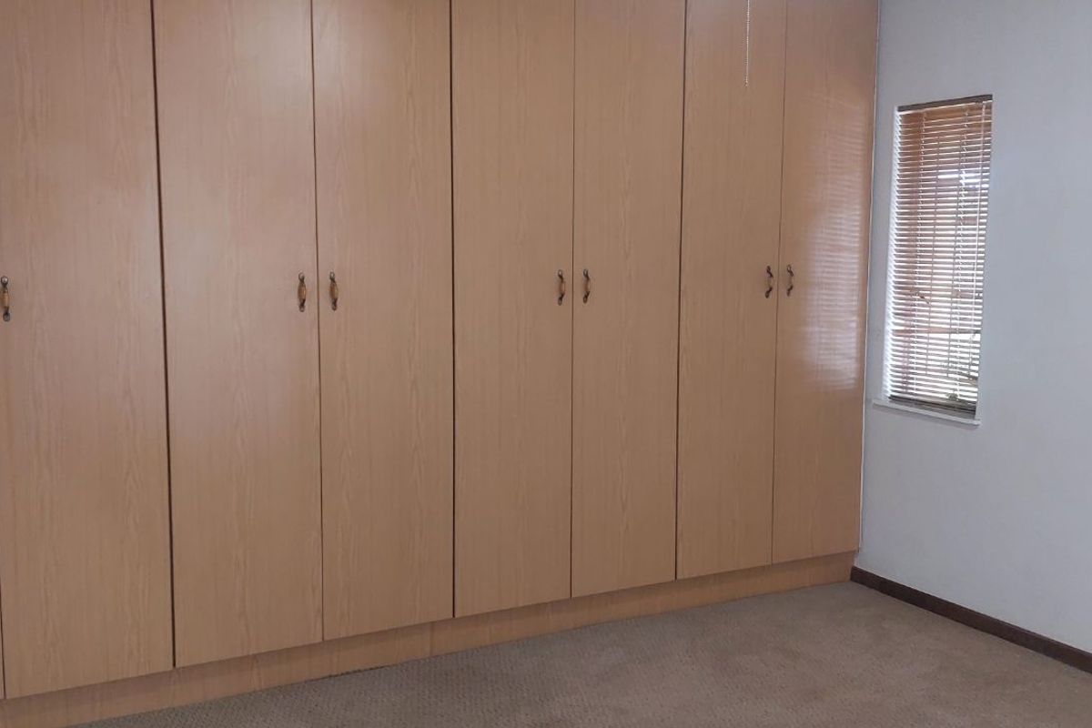 A bedroom featuring extensive wall-to-wall built-in wooden cabinetry, neutral carpeting, and a window fitted with horizontal blinds.