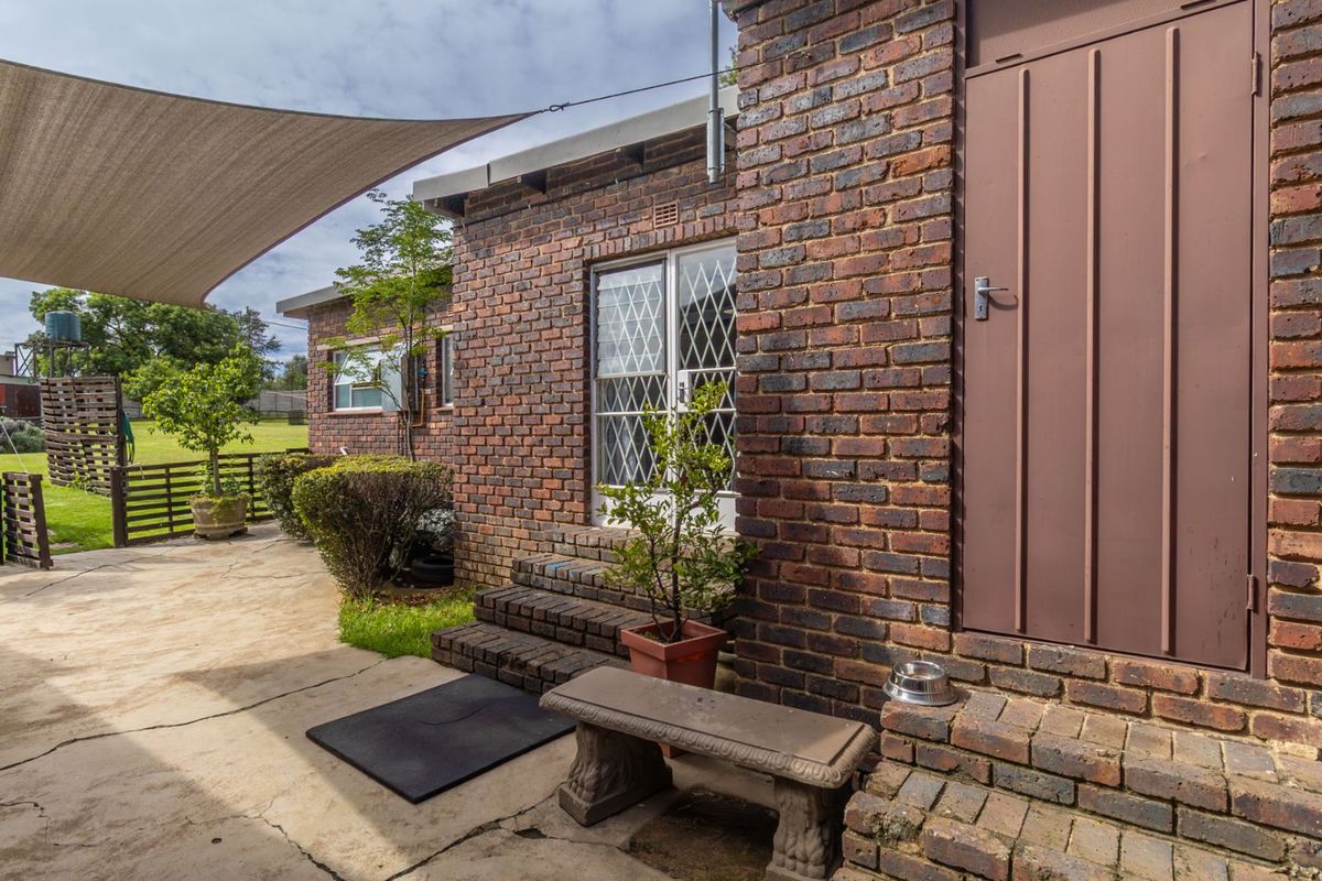 Rear exterior view of a brick farmhouse featuring a concrete patio, shade sail, and secure metal security gate entrance.