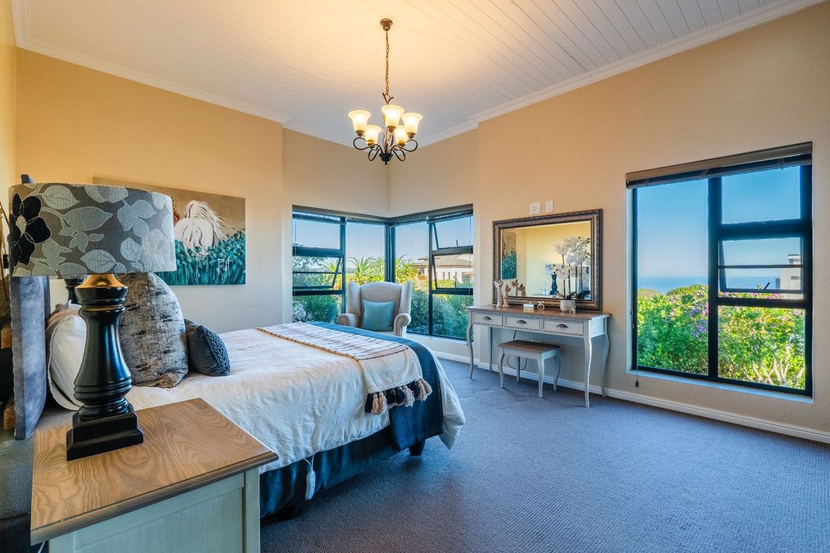 Spacious bedroom featuring large corner windows with ocean views, neutral wall tones, carpeted flooring, and a classic chandelier.