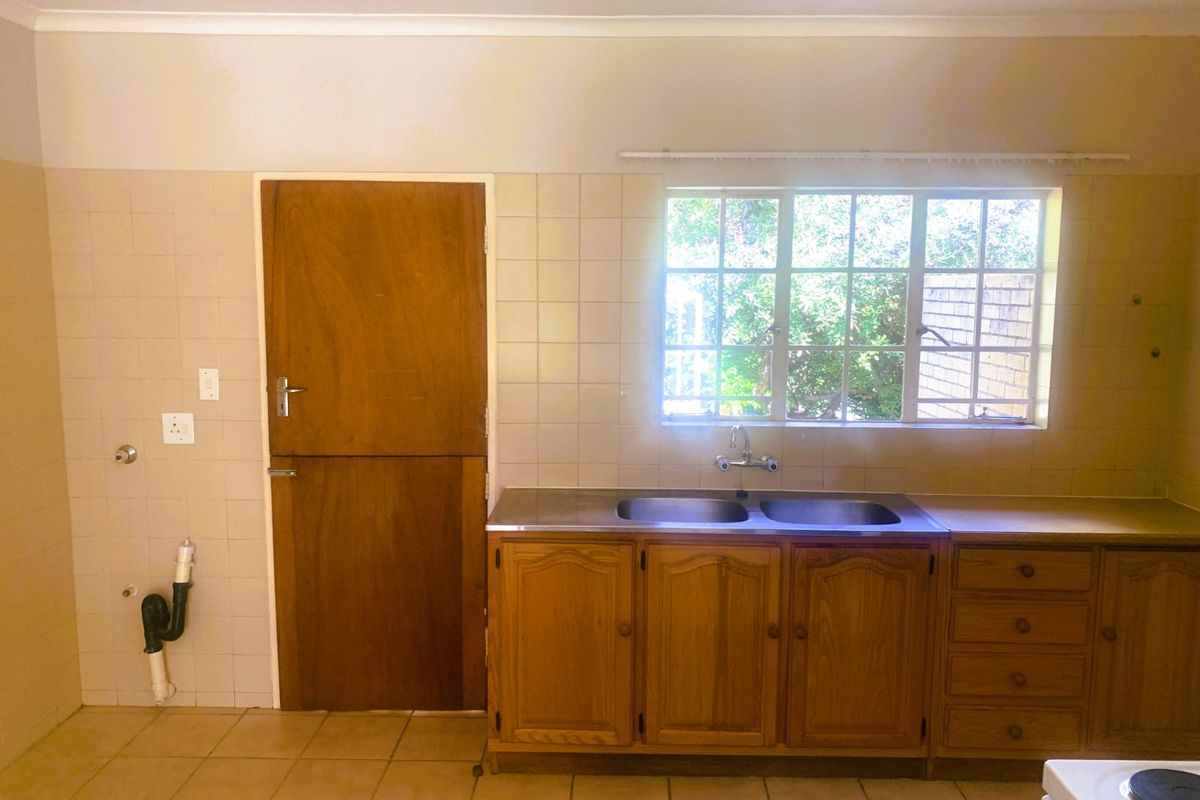 A dated kitchen area featuring wooden cabinetry, a double stainless steel sink, tiled splashbacks, and a window overlooking the garden.