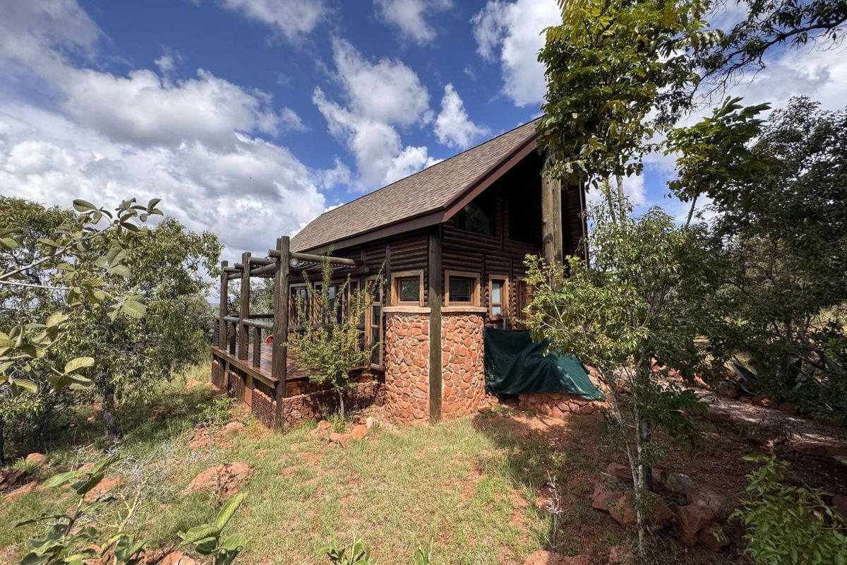A rustic log cabin style house featuring stone masonry accents, a wooden deck, and a pitched shingle roof set within a natural wooded landscape.