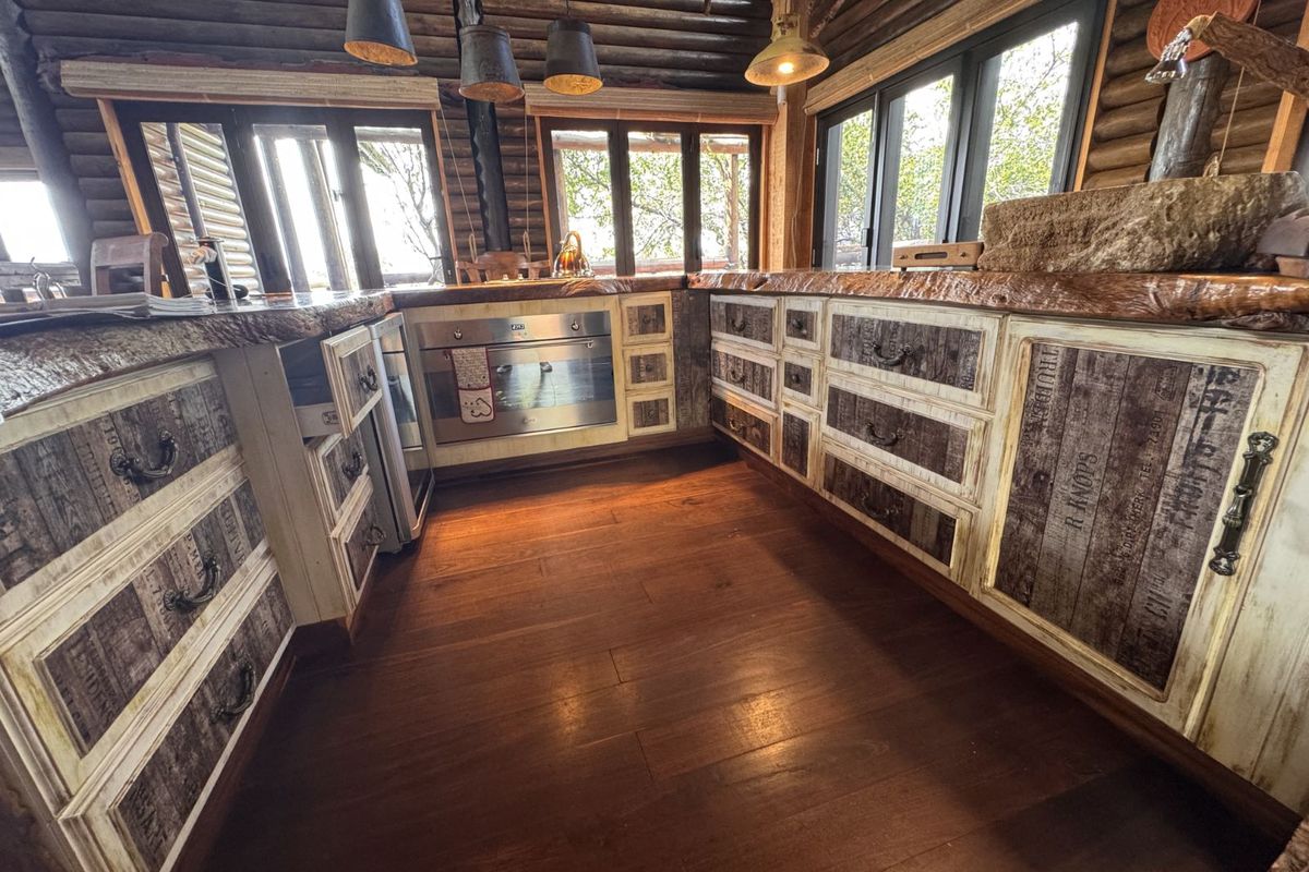 Rustic kitchen featuring custom timber cabinetry, live-edge wooden countertops, built-in oven, and exposed log walls with large windows providing natural light.