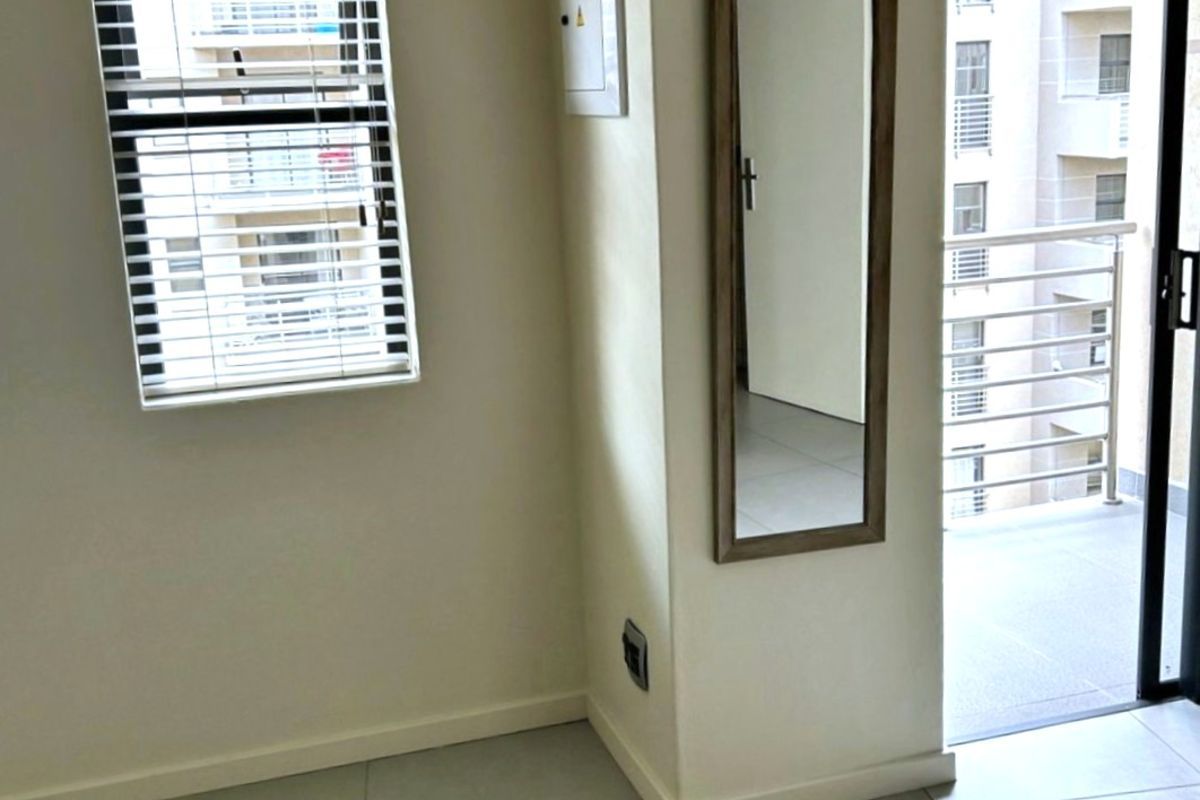 A bright living area featuring tiled flooring, a window with fitted blinds, and a sliding door leading to a private balcony.