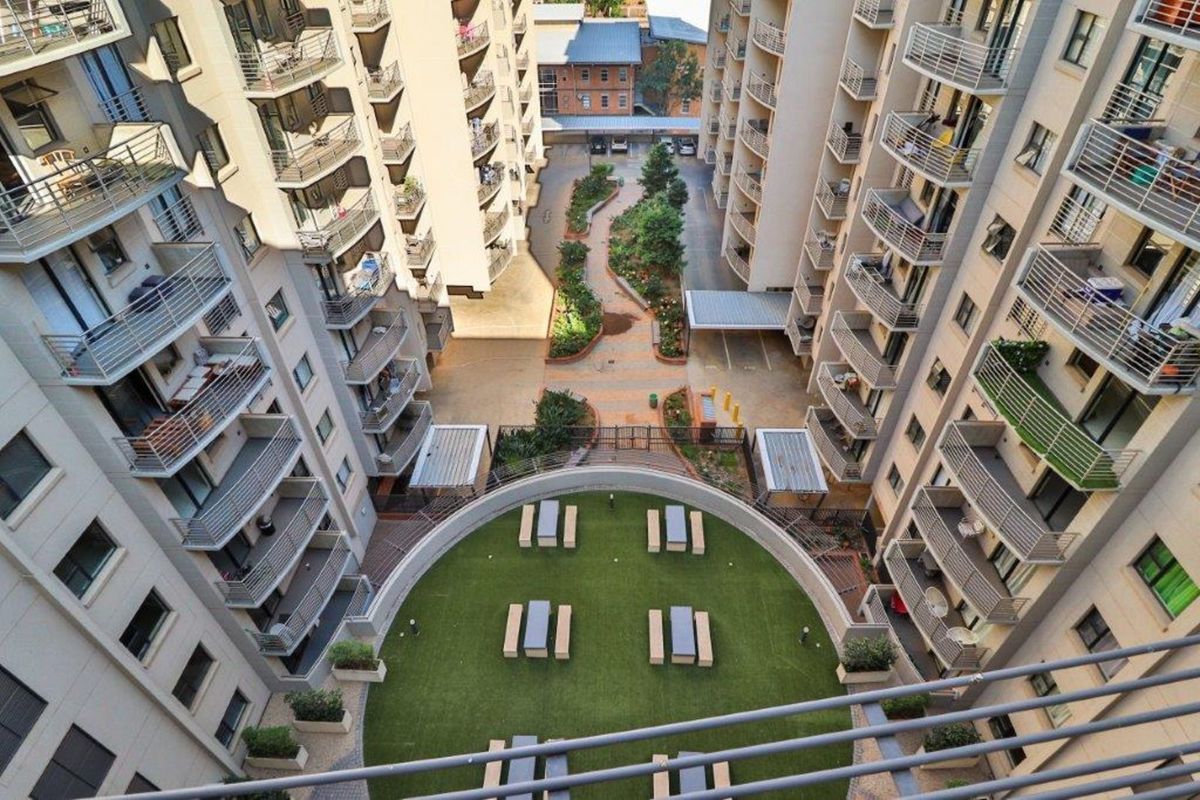 An elevated aerial view of an apartment complex courtyard featuring a landscaped green space, paved walkways, and covered parking areas.