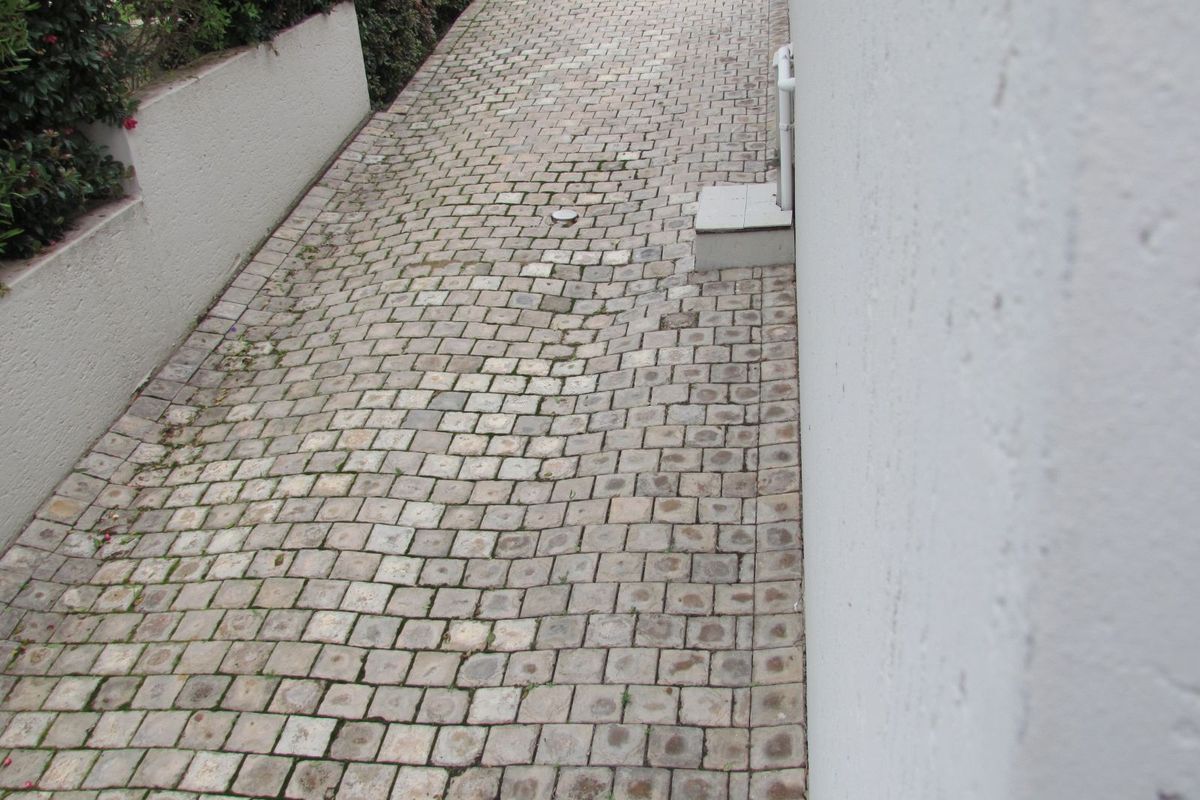 A narrow exterior side pathway featuring cobblestone paving, bordered by a low retaining wall and the side of the house structure.
