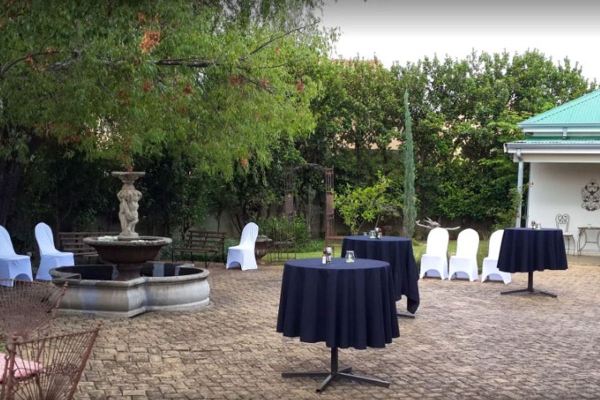 A paved outdoor courtyard area featuring a central stone fountain, mature trees, and a covered patio section attached to the main building structure.