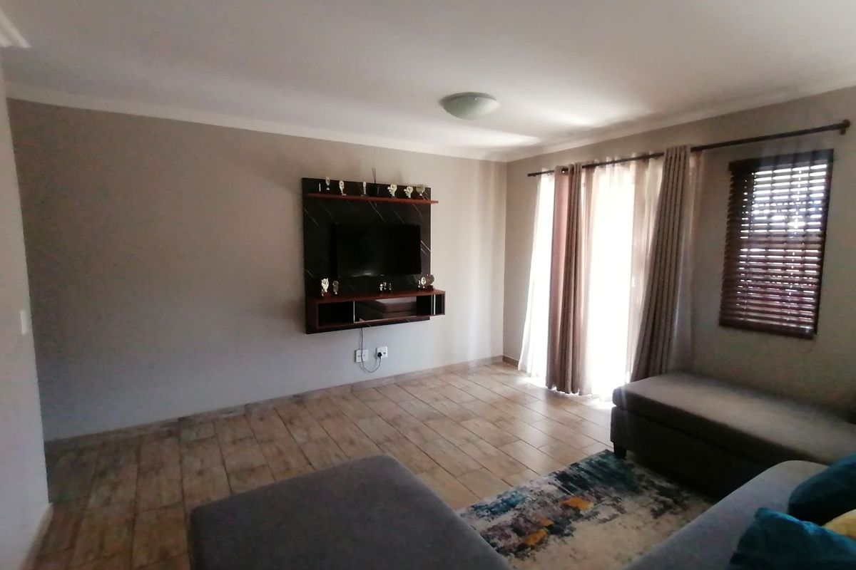 A well-maintained living room featuring tiled flooring, a built-in wall-mounted media unit, and large windows with blinds allowing for natural light.