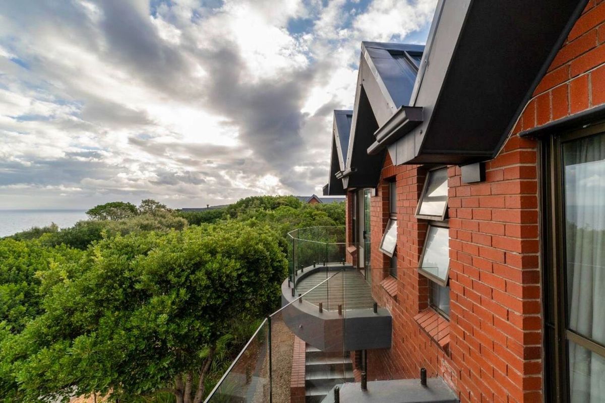 A view of the exterior rear facade featuring a curved balcony with glass balustrades, brick masonry, and metal roofing overlooking lush greenery and the ocean.