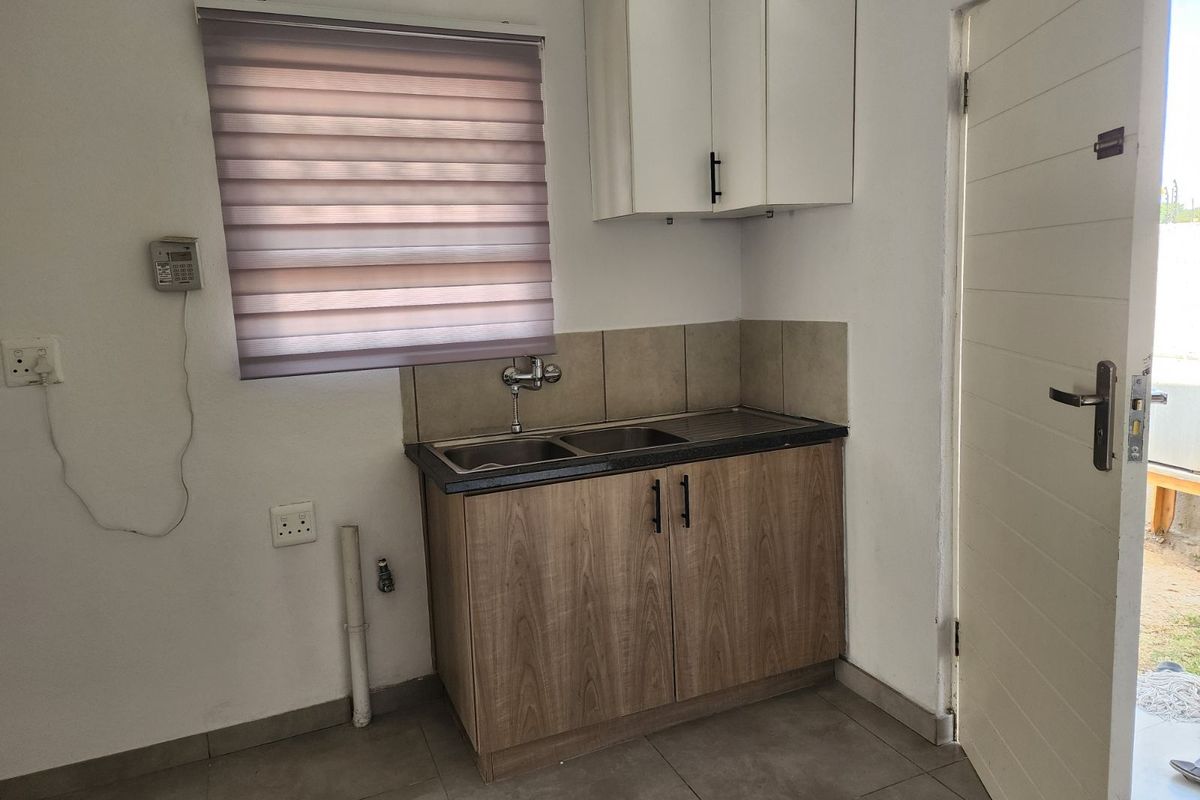 Compact kitchenette featuring wood-look cabinetry, a stainless steel double sink, tiled splashback, and a window with a roller blind.
