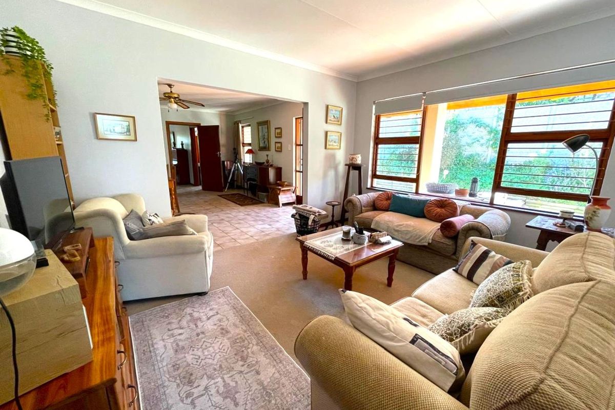 A spacious living room featuring large windows with louvre shutters, neutral carpeting, and an open-plan layout leading into an adjacent tiled area.