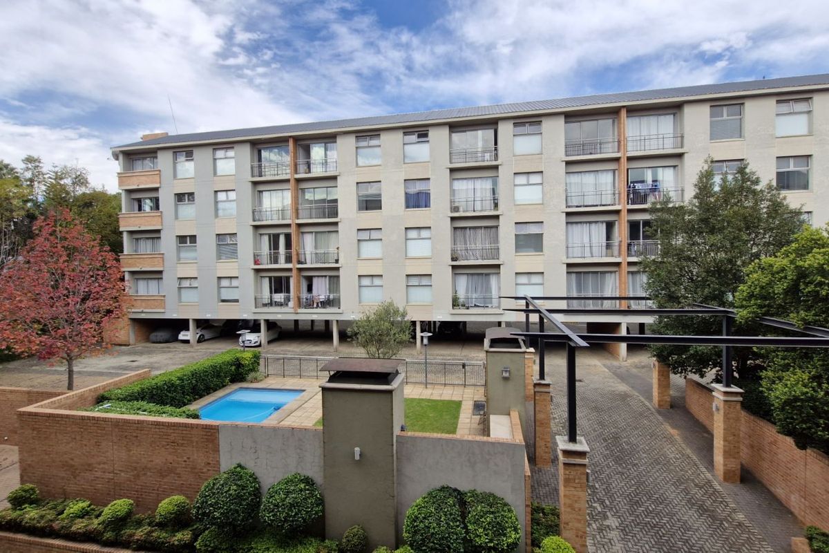 Exterior view of a multi-story apartment complex featuring a paved driveway, landscaped garden area, and a small swimming pool.