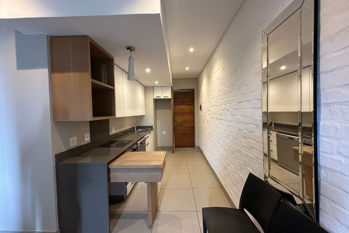 Modern galley-style kitchen featuring sleek cabinetry, integrated appliances, stone countertops, and a unique pull-out breakfast bar, complemented by a textured white brick feature wall.