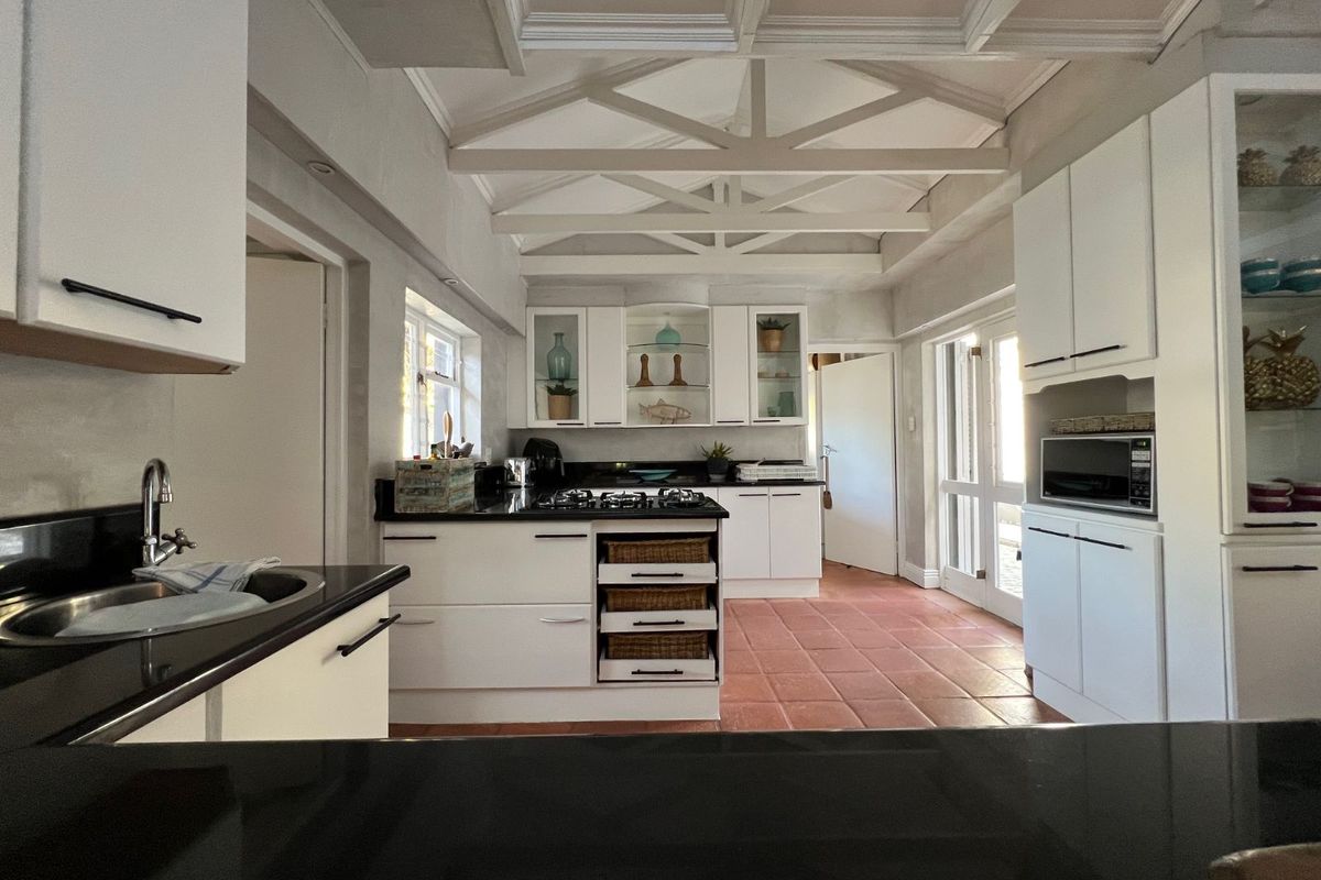 A bright, well-maintained kitchen featuring white cabinetry, black granite countertops, a gas hob, and a distinctive exposed beam ceiling with terracotta flooring.