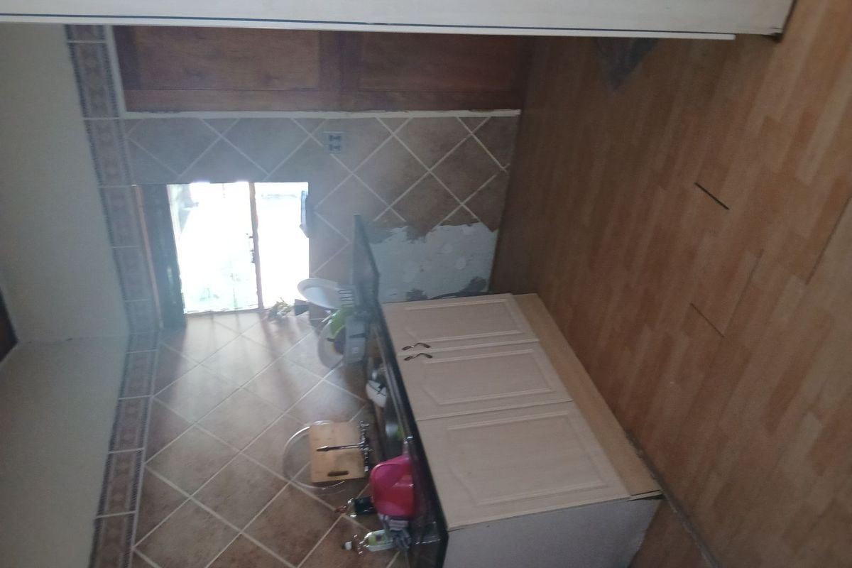 A small kitchen area featuring built-in cabinetry and tiled splashbacks, currently showing signs of wear and requiring maintenance to the wall and flooring.