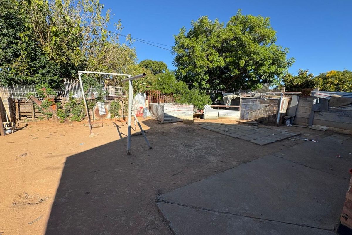 A spacious rear yard area featuring a mix of concrete paving and dirt ground, with existing metal structures and mature trees.