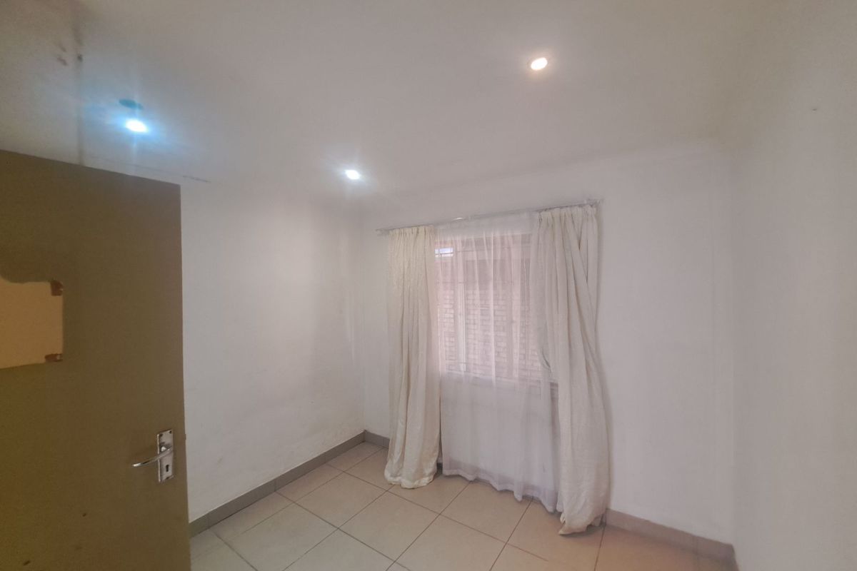 A simple, empty bedroom featuring tiled flooring, recessed ceiling lighting, and a window with curtain tracks.