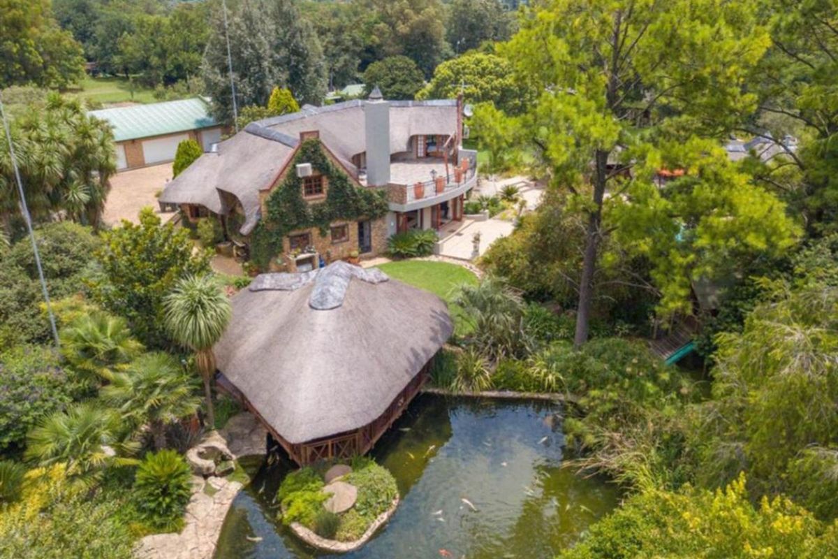 Aerial view of a property featuring a thatched-roof main house, a separate thatched gazebo over a koi pond, and lush landscaped gardens.