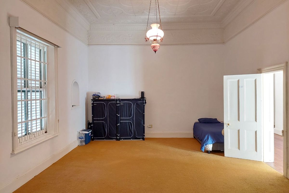 Spacious bedroom featuring high ceilings with decorative cornicing, a large window with security bars, and original architectural character.