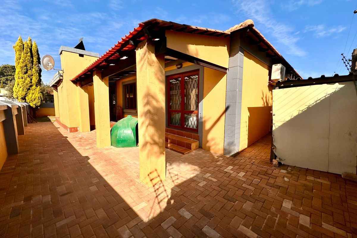 A rear exterior view of the townhouse featuring a paved patio area, covered veranda with support pillars, and secure access doors.