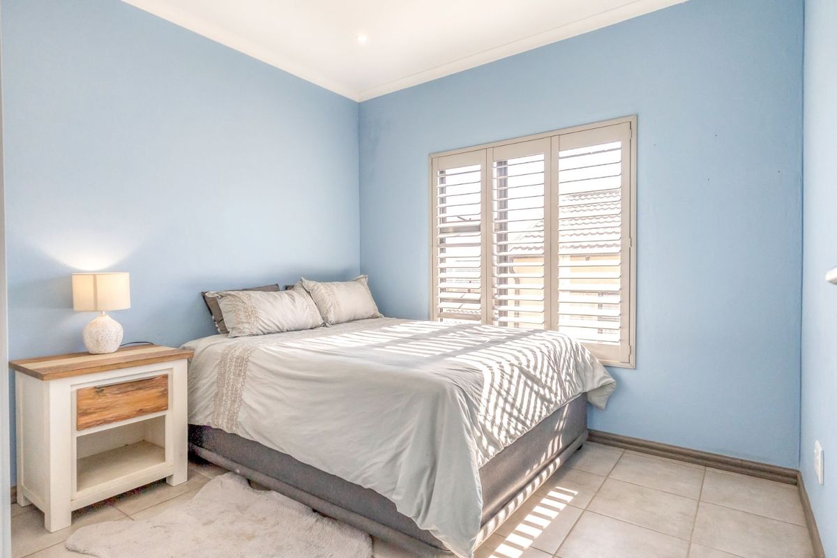 A bright bedroom featuring light blue walls, neutral tiled flooring, and fitted plantation shutters providing natural light control.