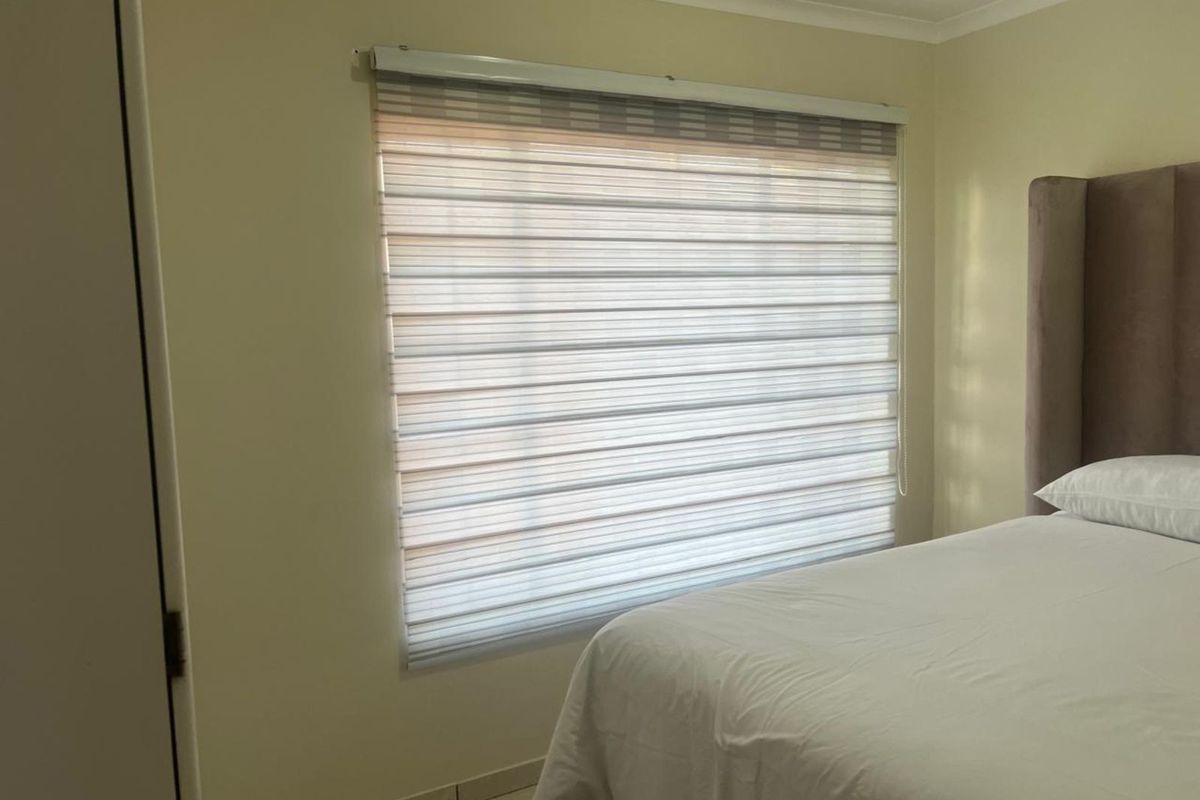 A well-maintained bedroom featuring neutral wall tones, tiled flooring, and modern zebra-style roller blinds for adjustable natural light control.