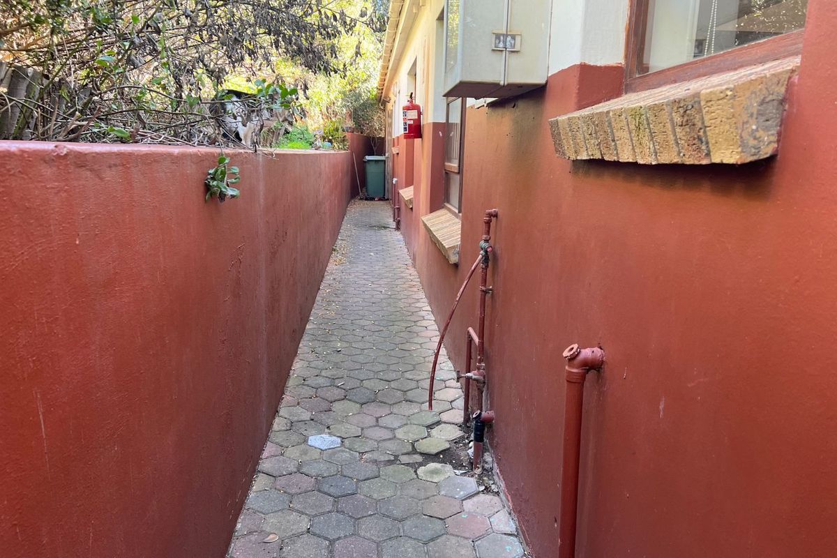 A narrow exterior side passage featuring paved flooring, boundary walls, and exposed external plumbing fixtures along the building facade.