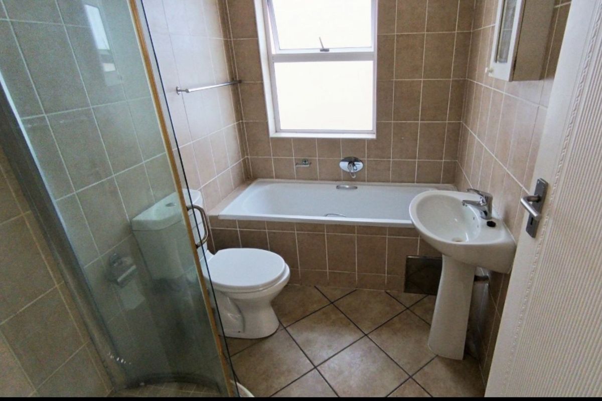 A functional bathroom featuring a tiled bathtub, pedestal basin, toilet, and a glass-enclosed shower with neutral wall and floor tiling.
