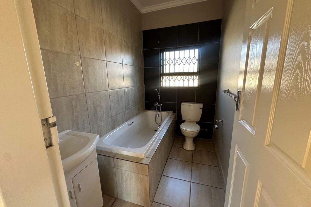 A functional bathroom featuring a built-in bathtub, pedestal basin, and toilet, finished with neutral wall and floor tiling and security bars on the window.