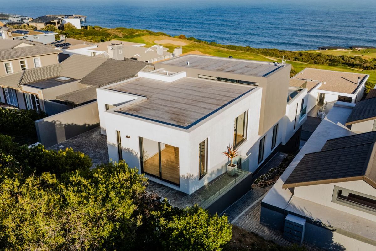 Aerial view of a modern coastal residence featuring a flat roof design, paved driveway, and glass balcony railings, situated near the ocean.