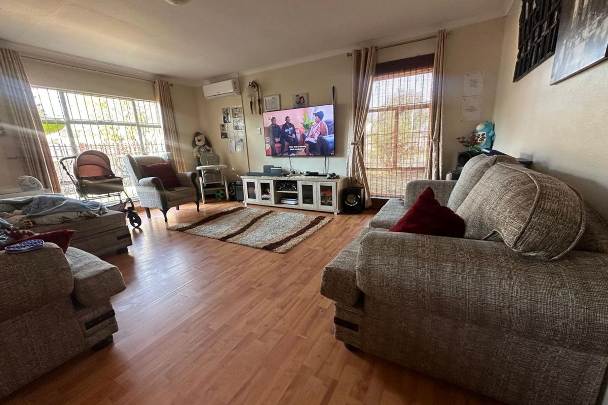 A spacious living room featuring wood-look laminate flooring, large windows with security bars, and a wall-mounted air conditioning unit.