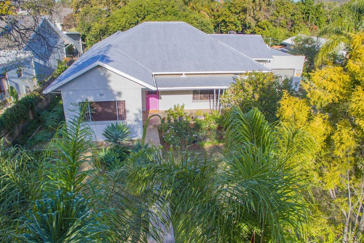 An elevated aerial view of a residential house featuring a pitched metal roof, front garden, and established trees.
