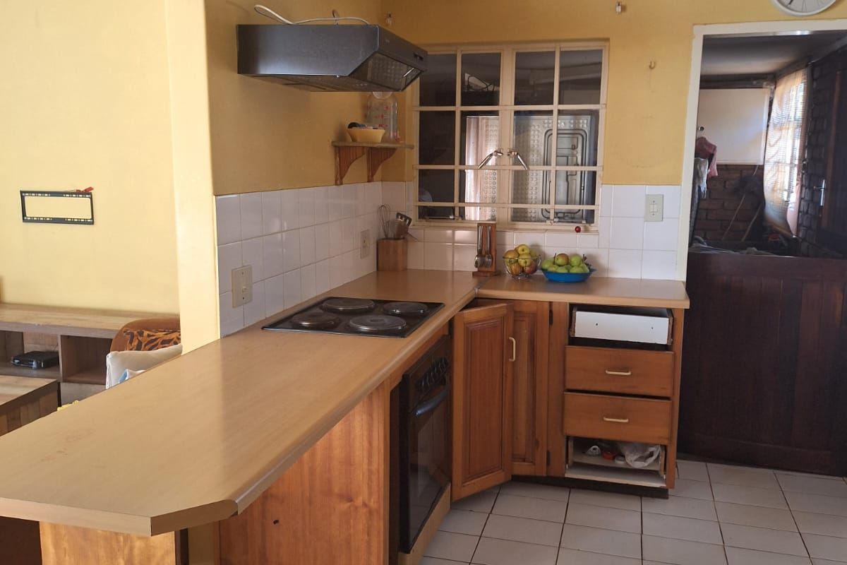 A dated kitchen featuring wooden cabinetry, a built-in hob and oven, tiled flooring, and a breakfast bar counter, opening into an adjacent living space.