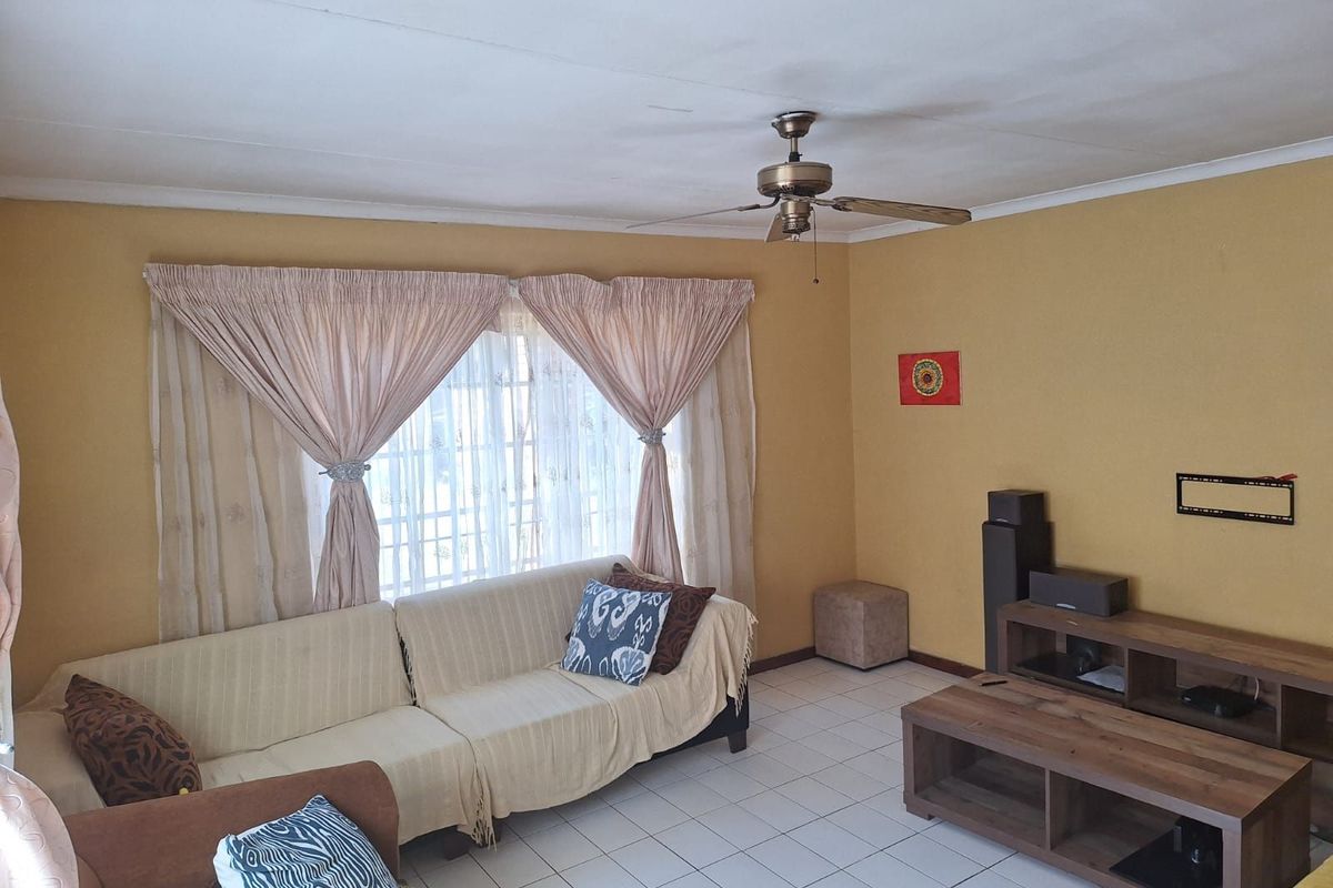 A living room featuring tiled flooring, a ceiling fan, and large windows with curtains, providing a functional and comfortable living space.