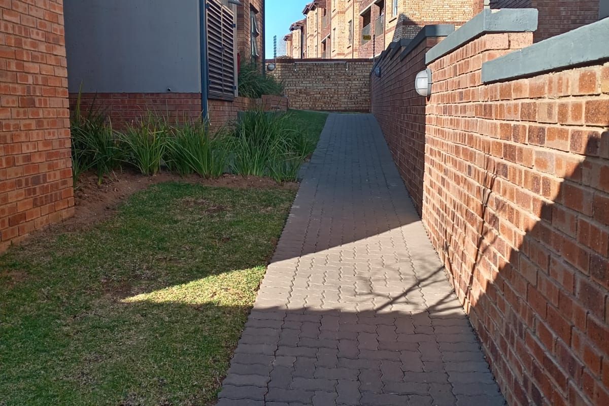 A paved walkway leading between apartment buildings, featuring brick exterior walls, landscaped garden patches, and exterior wall-mounted lighting.