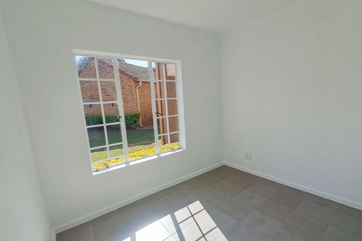 A bright, empty bedroom featuring neutral tiled flooring and a large window with a view of the exterior garden area.