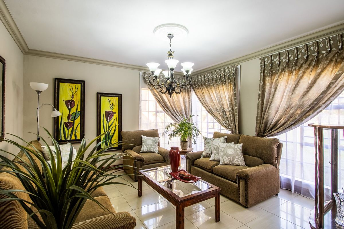 A well-lit living room featuring tiled flooring, decorative ceiling rose with a chandelier, and large windows with curtain treatments.