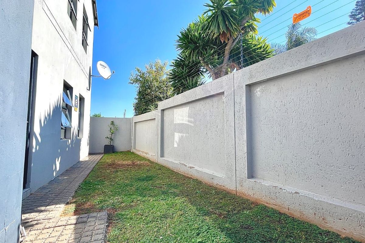 A side garden area featuring a paved walkway, a small lawn, and a high perimeter boundary wall with electric fencing.